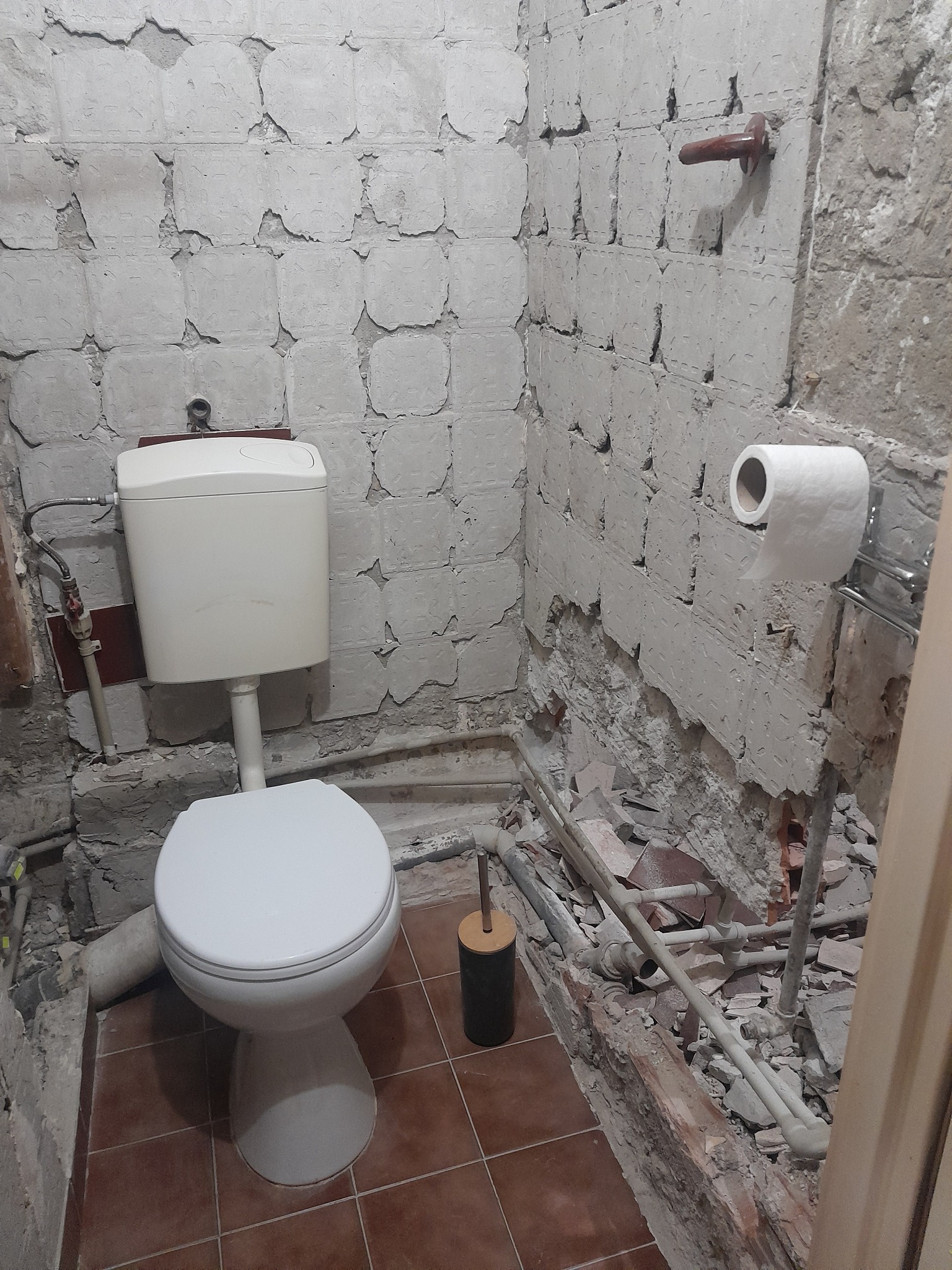 Demontaż płytek w łazience z widocznym WC, rurami i fragmentami gruzu. Ściany częściowo bez okładziny, widoczne instalacje. Na ścianie uchwyt na papier toaletowy.