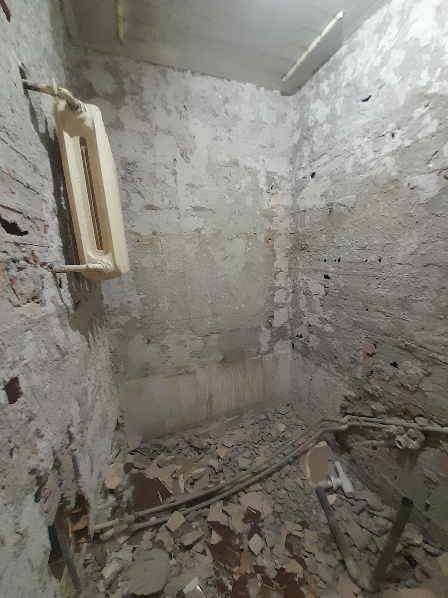 Wnętrze łazienki w trakcie demontażu: odsłonięte cegły, resztki gruzu na podłodze, stary grzejnik na ścianie. Widoczne rury instalacji wodnej.