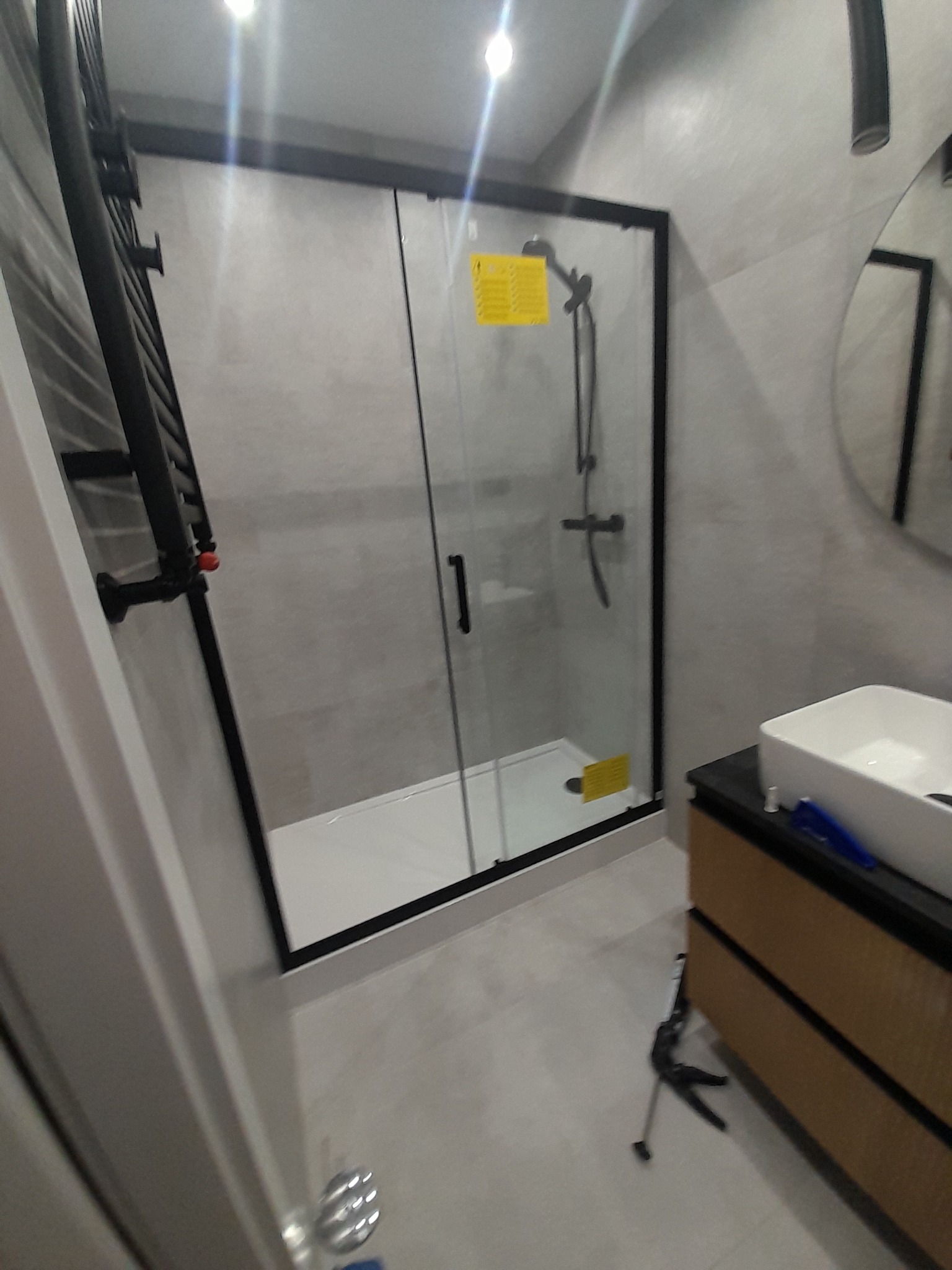 Nowoczesna łazienka po remoncie: kabina prysznicowa z czarną ramą, grzejnik drabinkowy i umywalka nablatowa. Minimalistyczny design, szare płytki.