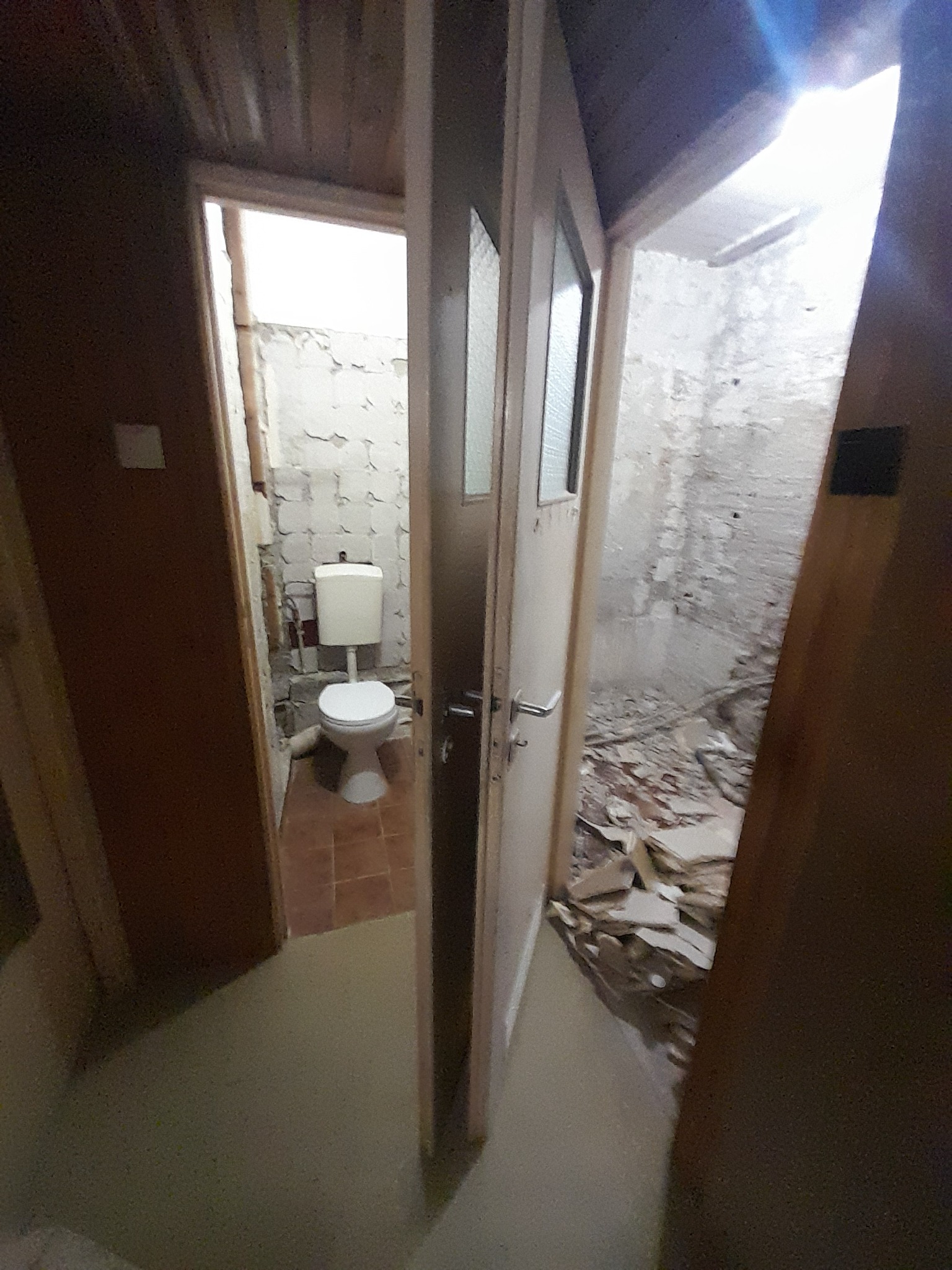 Łazienka w trakcie demontażu: widok przez otwarte drzwi na toaletę i zdemolowane ściany. Gruz na podłodze, odsłonięte instalacje. Etap przygotowawczy do remontu.