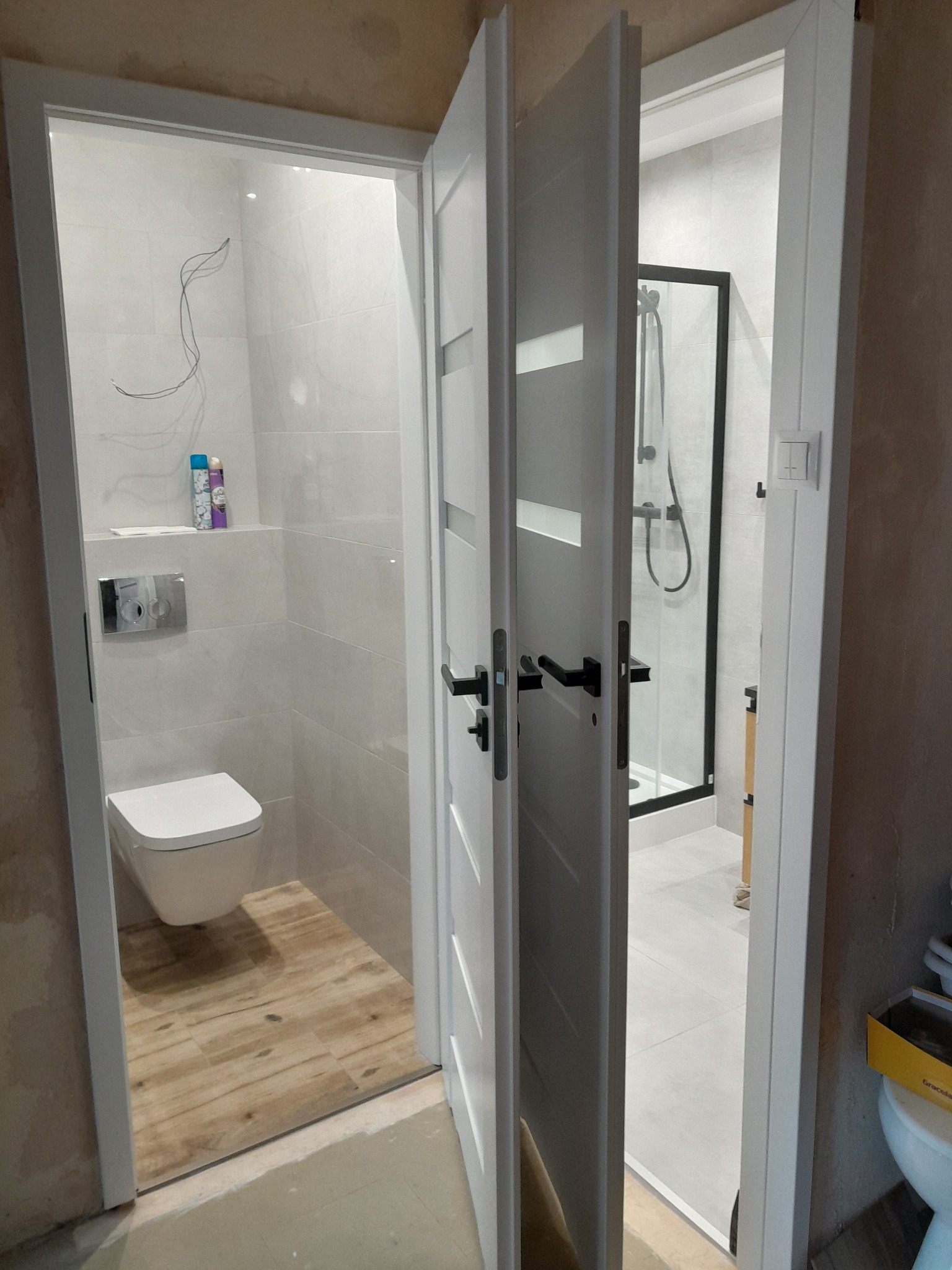 Dwa wejścia do łazienki z białymi drzwiami. W jednej toaleta z drewnopodobną podłogą, w drugiej kabina prysznicowa z czarną ramą. Ściany wyłożone jasnymi płytkami.