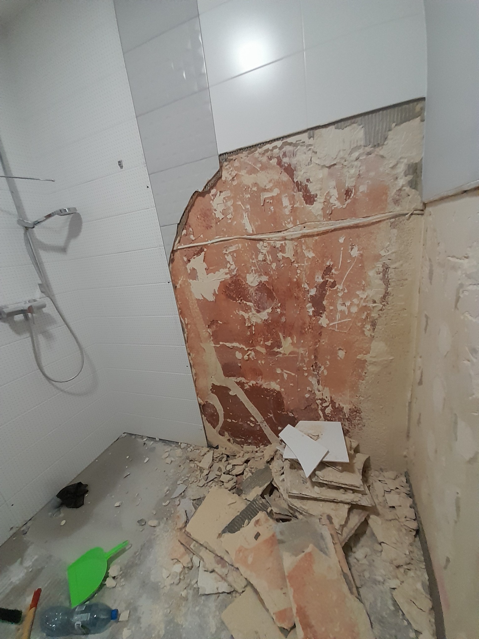 Demontaż płytek w łazience: fragment ściany z resztkami kleju, odspojone płytki na podłodze, widoczny prysznic i narzędzia. Etap przygotowawczy do remontu.