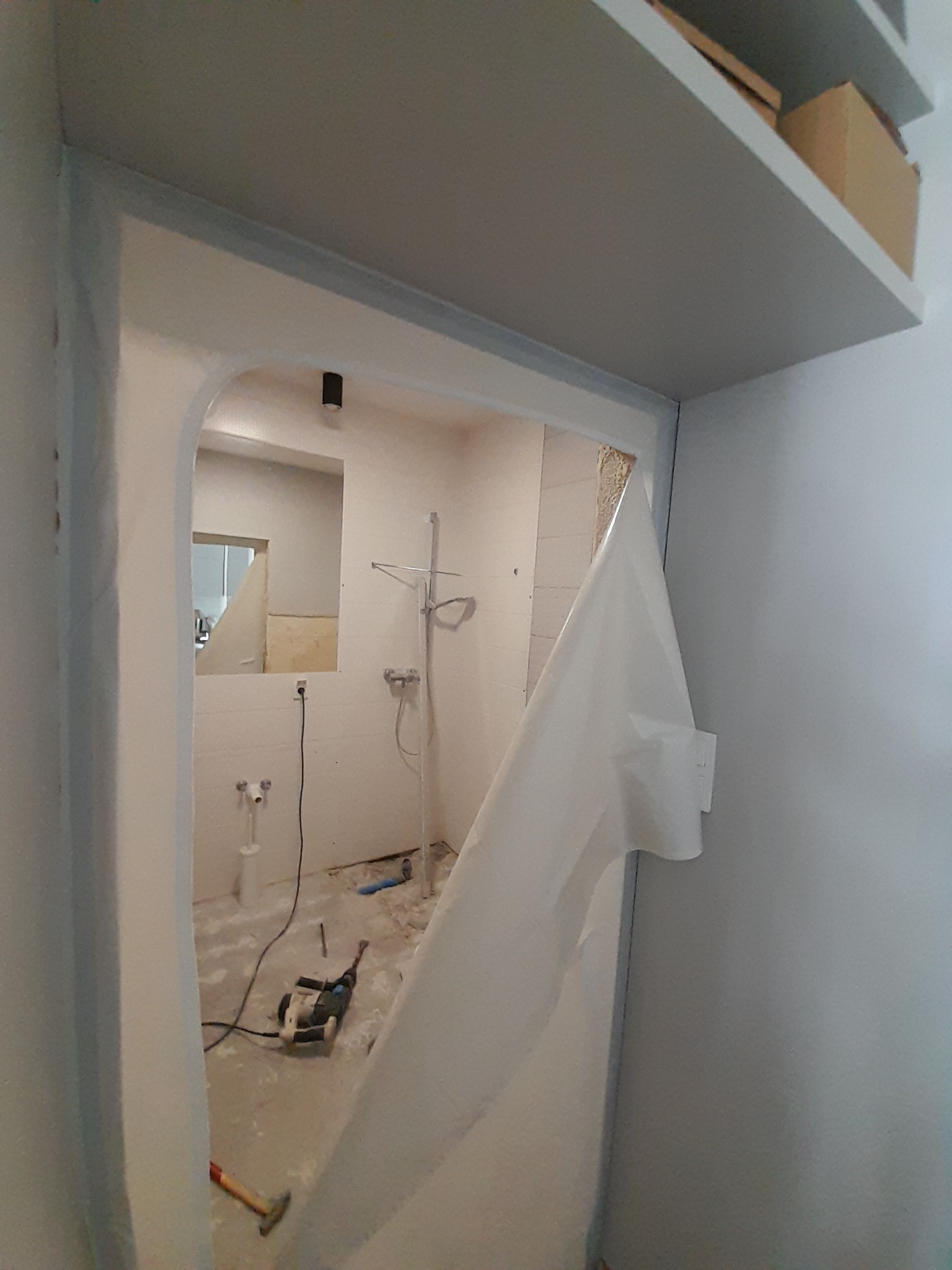 Wnętrze łazienki w trakcie demontażu, widoczne narzędzia, fragment instalacji i zabezpieczająca folia. Ujęcie z perspektywy wejścia, z półką w tle.