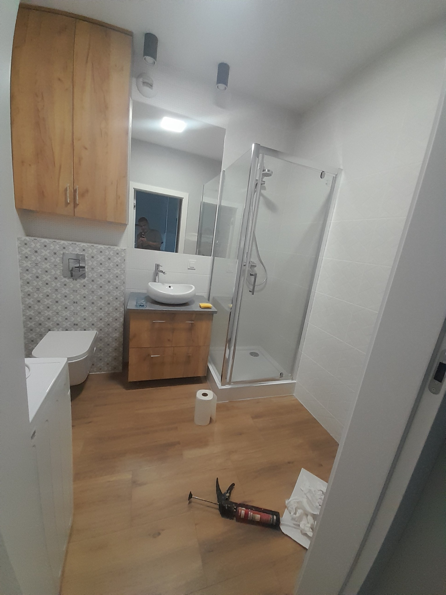 Jasna łazienka po remoncie: kabina prysznicowa, umywalka z szafką, toaleta. Na podłodze narzędzia i papier. Widok zza futryny.