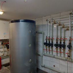 SMART INSTAL MARIUSZ KUBASIEWICZ - Instalacja centralnego ogrzewania w kotłowni z dwoma białymi piecami, szarym zasobnikiem ciepłej wody, czerwonym naczyniem przeponowym i rozbudowaną siecią rur z zaworami na tle białych płytek.