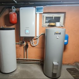 SMART INSTAL MARIUSZ KUBASIEWICZ - Instalacja grzewcza w piwnicy: biały zbiornik, czerwony zbiornik wyrównawczy i szary zbiornik Ferroli, połączone rurami na tle pomarańczowej ściany. Dodatkowo widoczna szafka elektryczna i okno.