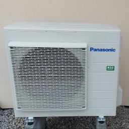 SMART INSTAL MARIUSZ KUBASIEWICZ - Zewnętrzna jednostka klimatyzacji Panasonic na betonowych podstawach, widoczne krople wody na obudowie i logo R32 refrigerant.