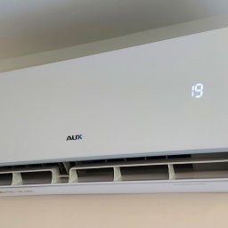 SMART INSTAL MARIUSZ KUBASIEWICZ - Biała jednostka klimatyzacji marki AUX zamontowana na beżowej ścianie, wyświetlająca temperaturę 19 stopni Celsjusza.