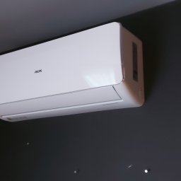 SMART INSTAL MARIUSZ KUBASIEWICZ - Biała jednostka wewnętrzna klimatyzatora marki AUX zamontowana na ciemnoszarej ścianie, widoczne drobne punkty światła odbijające się od ściany, fragment pomarańczowej zasłony w tle.