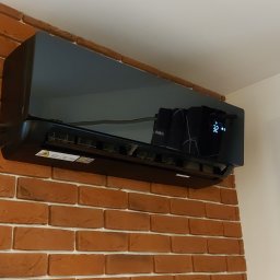 SMART INSTAL MARIUSZ KUBASIEWICZ - Czarny klimatyzator zamontowany na ścianie z cegły dekoracyjnej, widoczne detale: wyświetlacz z temperaturą 32 stopnie, naklejki ostrzegawcze i fragment zasłony z geometrycznym wzorem.