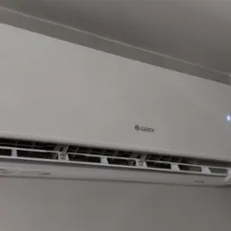 Biała jednostka klimatyzacji marki Gree zamontowana na szarej ścianie, wyświetlająca temperaturę 16 stopni Celsjusza.