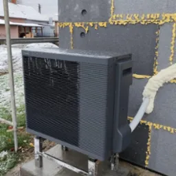 Szara jednostka zewnętrzna urządzenia grzewczego na chromowanych nóżkach, zamontowana na betonowym fundamencie przy ścianie z szarych bloczków z żółtą pianką uszczelniającą, na tle zaśnieżonego...