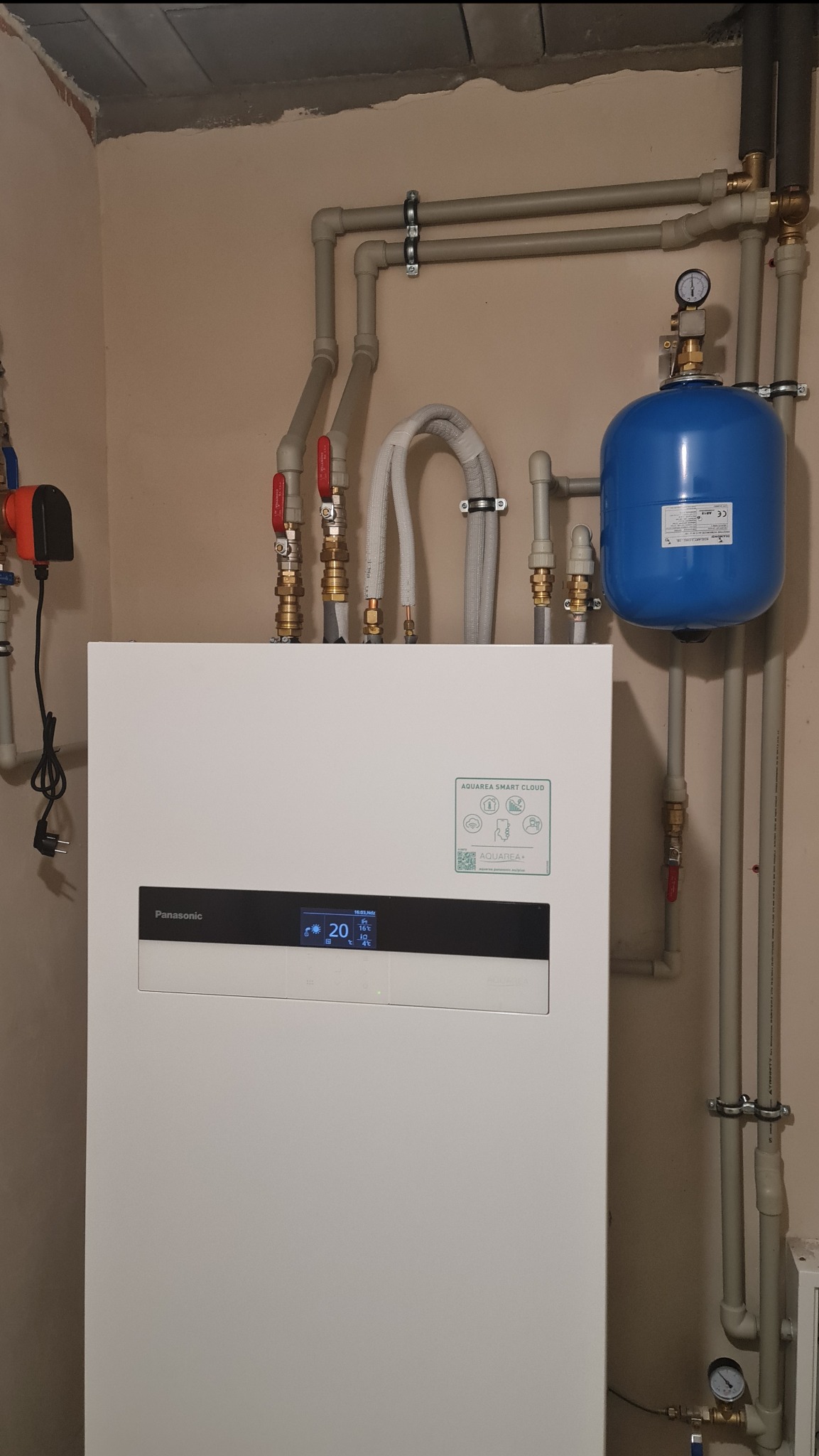 Biała pompa ciepła Panasonic Aquarea z niebieskim zbiornikiem wyrównawczym i instalacją hydrauliczną w pomieszczeniu w Zagajniku, widoczne rury i zawory.