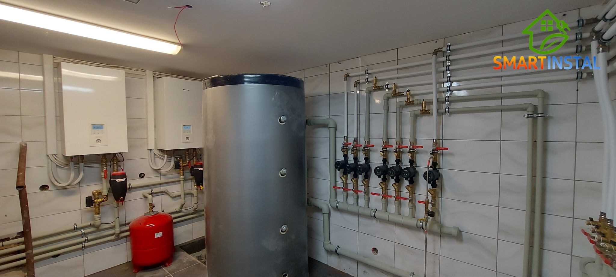 Instalacja centralnego ogrzewania w kotłowni z dwoma białymi piecami, szarym zasobnikiem ciepłej wody, czerwonym naczyniem przeponowym i rozbudowaną siecią rur z zaworami na tle białych płytek.