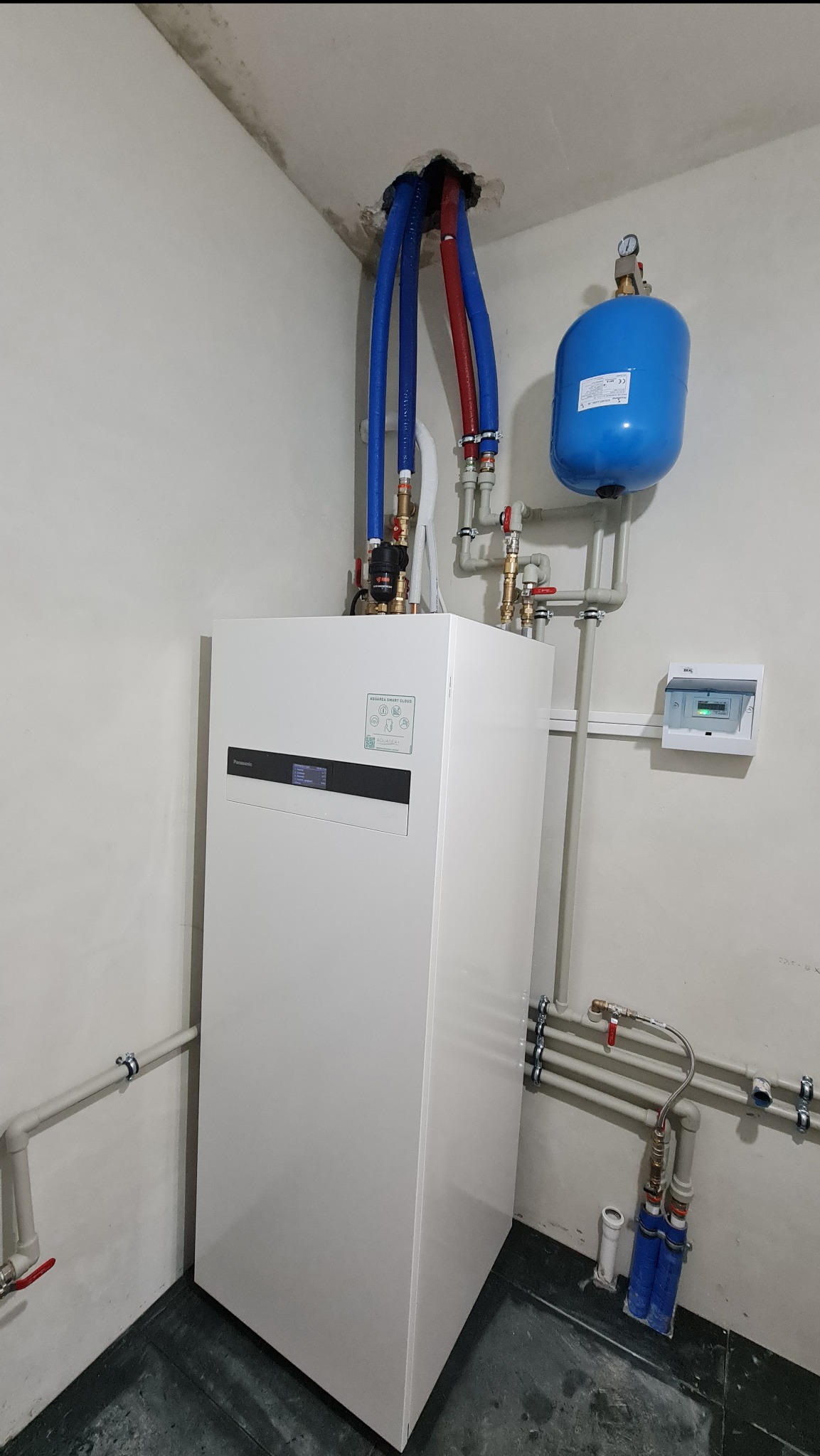 Biała pompa ciepła Panasonic Aquarea Smart Cloud zainstalowana w pomieszczeniu technicznym z widocznymi niebieskimi i czerwonymi rurami wychodzącymi z sufitu oraz osprzętem hydraulicznym.
