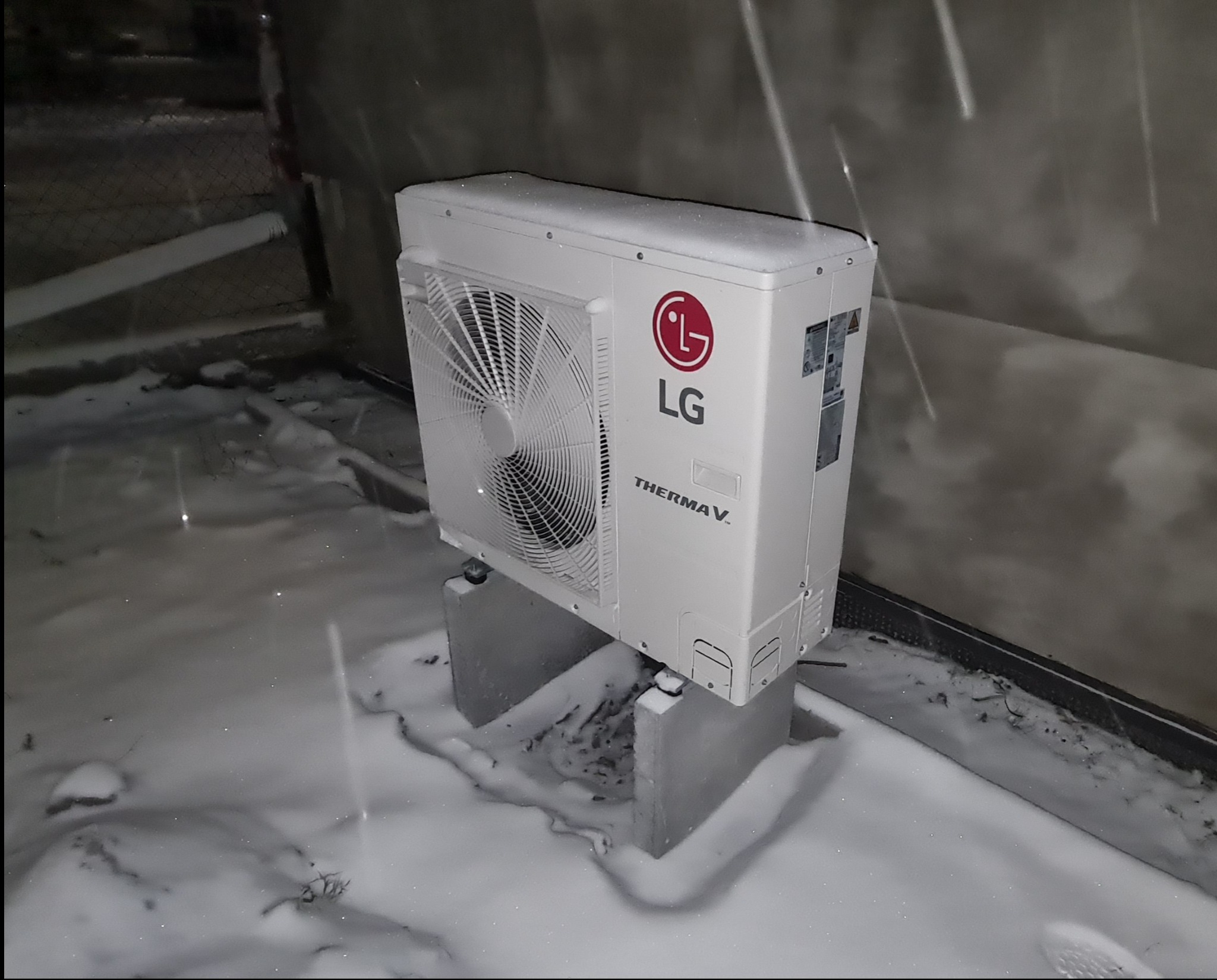 Biała zewnętrzna jednostka pompy ciepła LG Therma V, umieszczona na betonowych wspornikach, otoczona śniegiem, widok z boku.