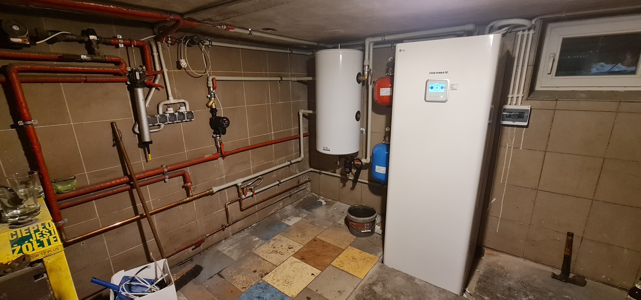 Kotłownia z zainstalowaną białą pompą ciepła LG Therma V, zbiornikiem buforowym, licznymi rurami miedzianymi pomalowanymi na czerwono, oraz fragmentem okna i skrzynki elektrycznej na ścianie...