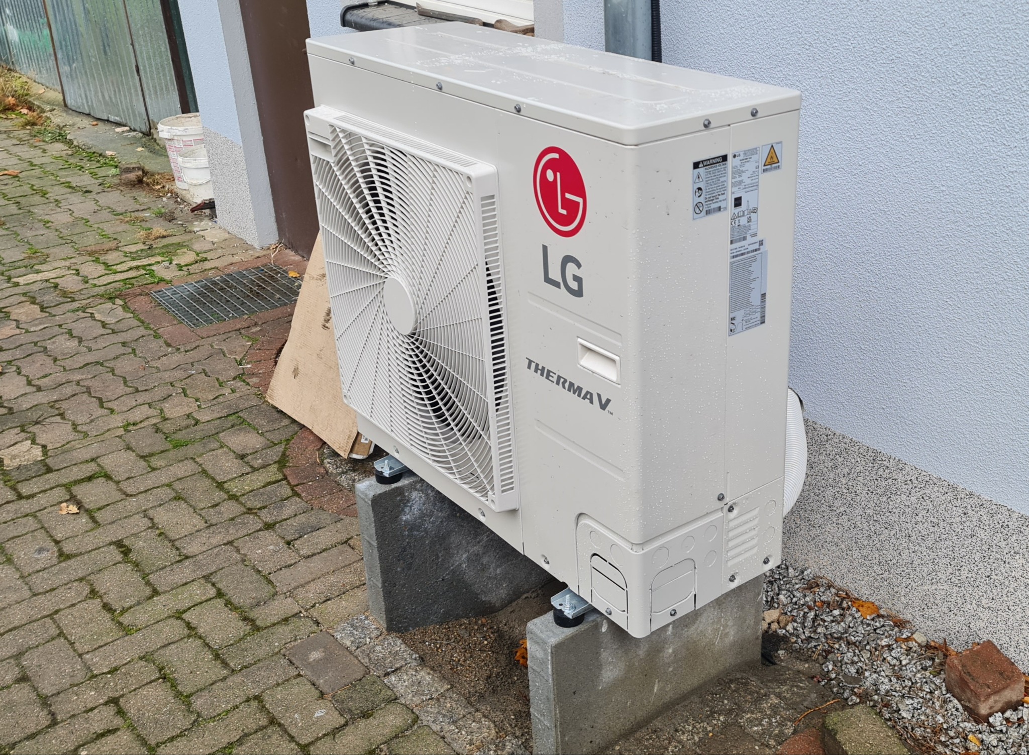 Zewnętrzna jednostka pompy ciepła LG Therma V, ustawiona na betonowych blokach przy ścianie budynku z szarą elewacją i brukowanym podłożem, widoczne krople deszczu na obudowie.