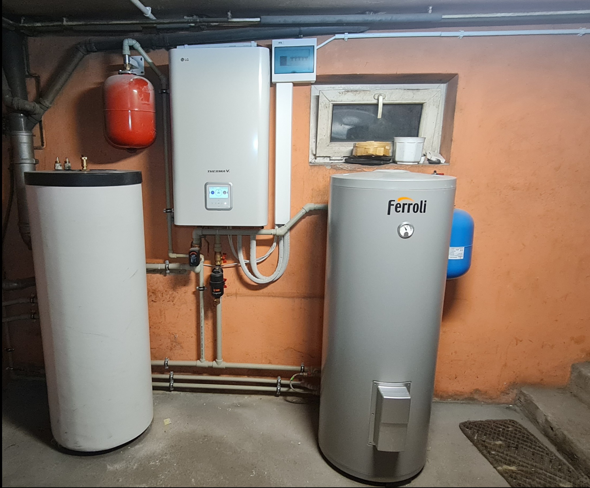 Instalacja grzewcza w piwnicy: biały zbiornik, czerwony zbiornik wyrównawczy i szary zbiornik Ferroli, połączone rurami na tle pomarańczowej ściany. Dodatkowo widoczna szafka elektryczna i okno.