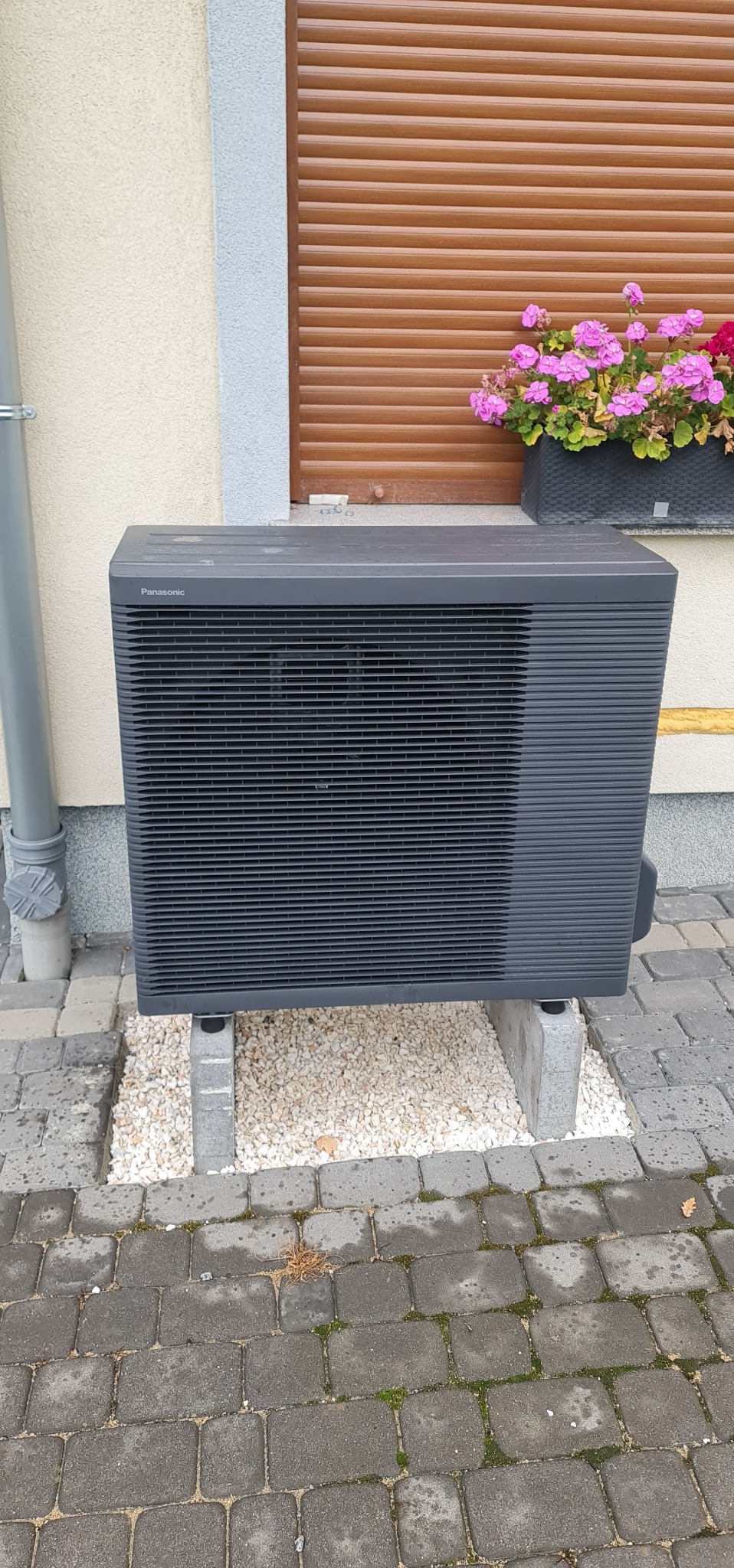 Zewnętrzna jednostka pompy ciepła Panasonic, umieszczona na betonowych bloczkach, z widocznym drenażem z białego żwiru, na tle ściany budynku z brązowymi roletami i skrzynką z różowymi kwiatami.