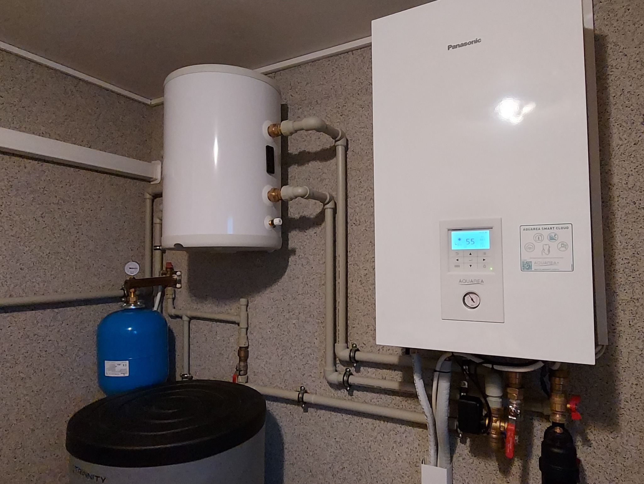 Biała pompa ciepła Panasonic Aquarea z podłączonym zasobnikiem ciepłej wody i niebieskim naczyniem przeponowym, zamontowana na ścianie o szarej, chropowatej fakturze.