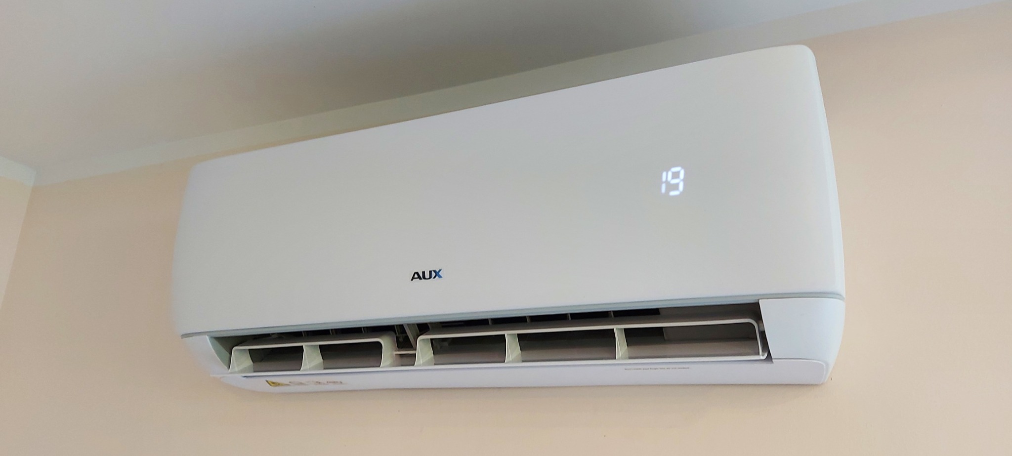 Biała jednostka klimatyzacji marki AUX zamontowana na beżowej ścianie, wyświetlająca temperaturę 19 stopni Celsjusza.