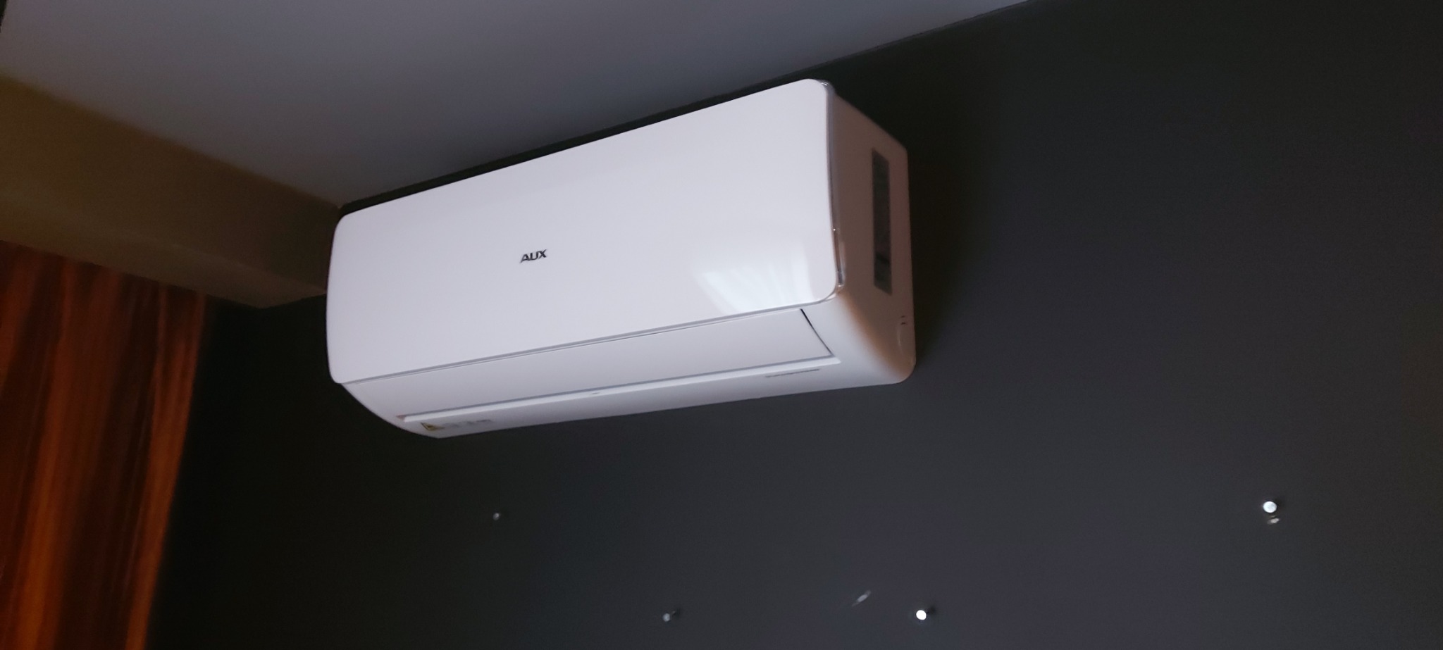 Biała jednostka wewnętrzna klimatyzatora marki AUX zamontowana na ciemnoszarej ścianie, widoczne drobne punkty światła odbijające się od ściany, fragment pomarańczowej zasłony w tle.