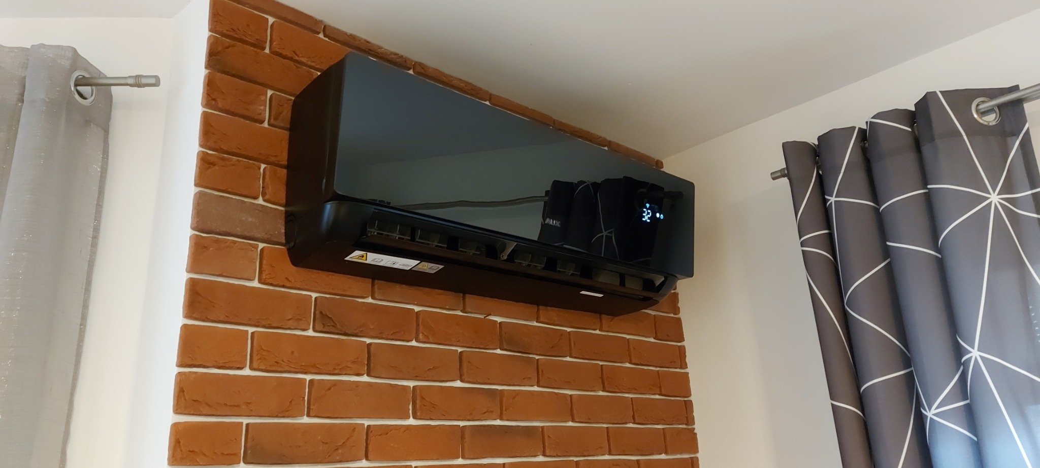 Czarny klimatyzator zamontowany na ścianie z cegły dekoracyjnej, widoczne detale: wyświetlacz z temperaturą 32 stopnie, naklejki ostrzegawcze i fragment zasłony z geometrycznym wzorem.