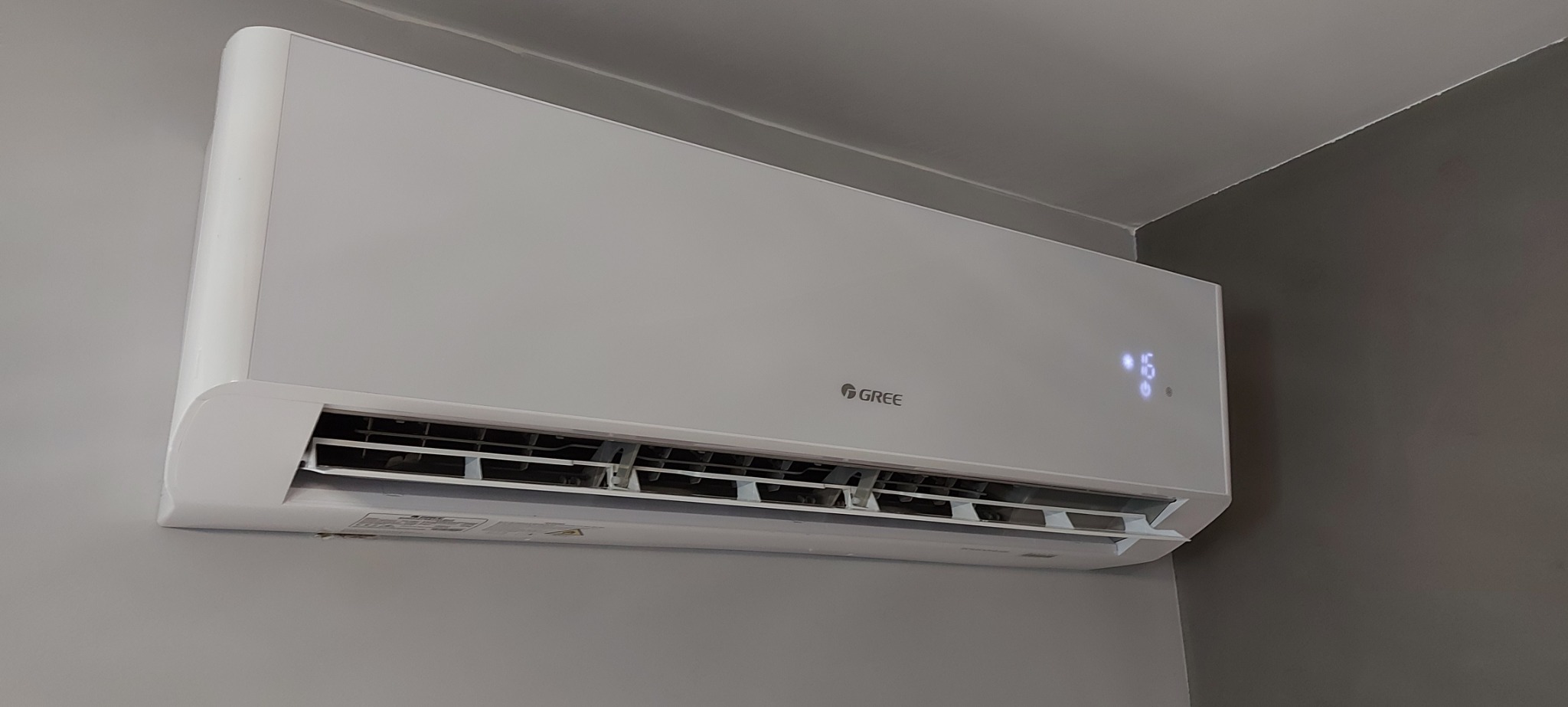 Biała jednostka klimatyzacji marki Gree zamontowana na szarej ścianie, wyświetlająca temperaturę 16 stopni Celsjusza.