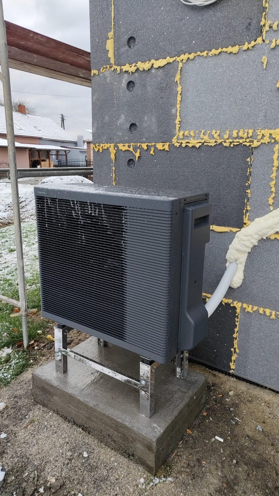 Szara jednostka zewnętrzna urządzenia grzewczego na chromowanych nóżkach, zamontowana na betonowym fundamencie przy ścianie z szarych bloczków z żółtą pianką uszczelniającą, na tle zaśnieżonego...