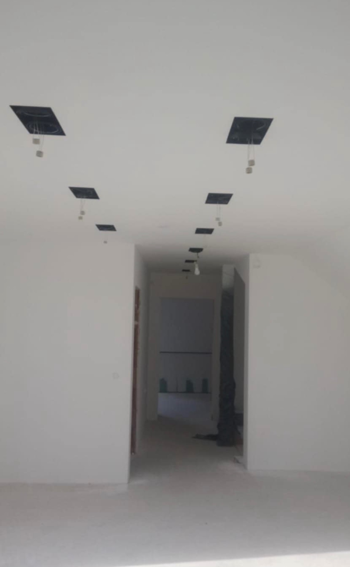 Wnętrze w trakcie remontu: białe ściany, surowy sufit z wyciętymi otworami na oświetlenie, widoczne kable i żarówki. Podłoga zabezpieczona folią malarską.