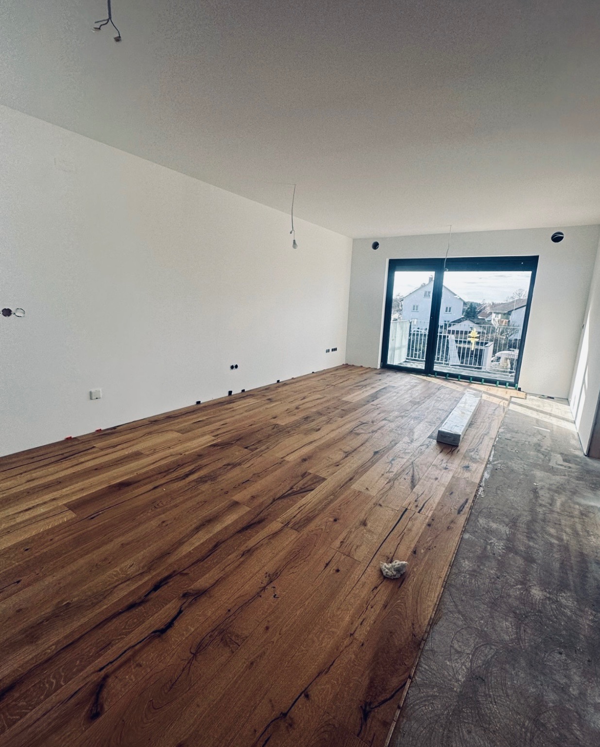 Wnętrze w trakcie układania podłogi z desek o ciepłym, brązowym odcieniu; widoczne niedokończone fragmenty podłoża, paczka desek oraz wystające kable elektryczne ze ściany.