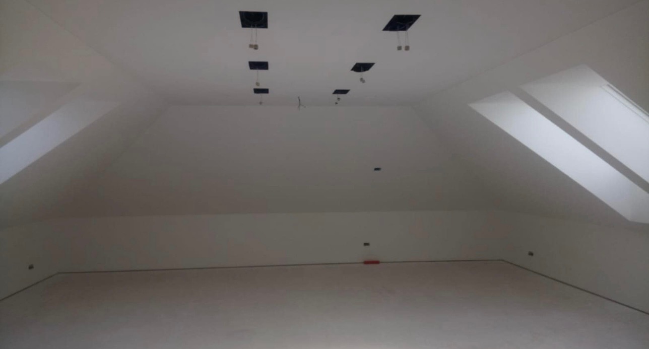 Poddasze w trakcie wykańczania, białe ściany i sufit, widoczne okna dachowe oraz otwory na oświetlenie sufitowe, brak umeblowania.