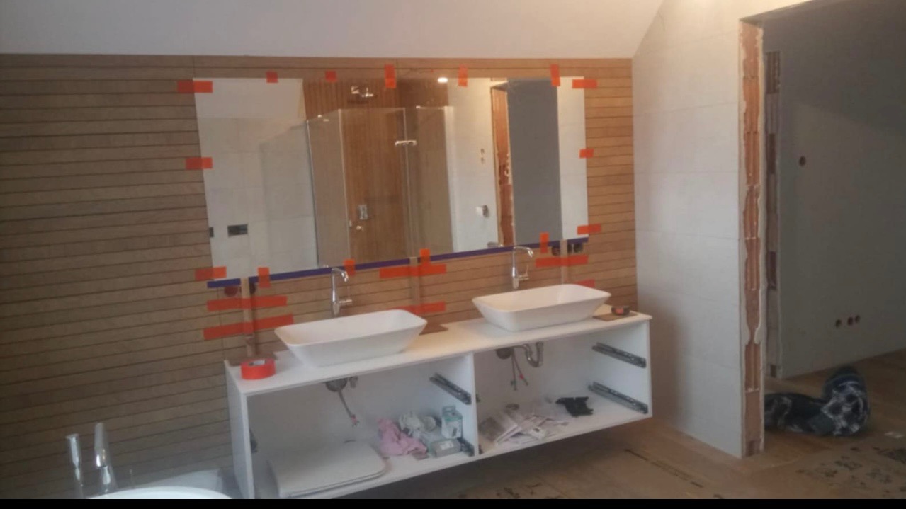 Łazienka w trakcie wykańczania: podwójna umywalka nablatowa na białej szafce, lustra przyklejone taśmą, drewnopodobne panele na ścianie, widoczne instalacje i narzędzia, surowy otwór drzwiowy.