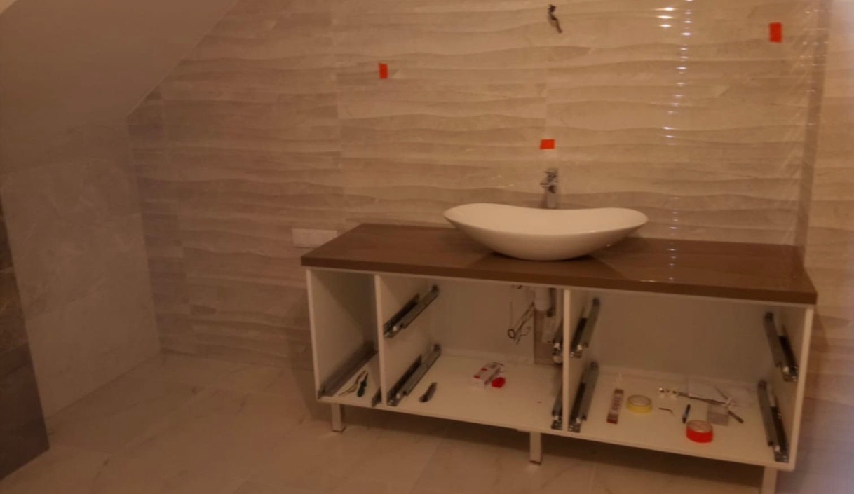 Łazienka w trakcie wykańczania: zamontowana szafka z blatem w kolorze drewna i owalną umywalką nablatową, widoczne elementy montażowe szuflad, ściany wyłożone beżowymi płytkami z delikatnym wzorem...