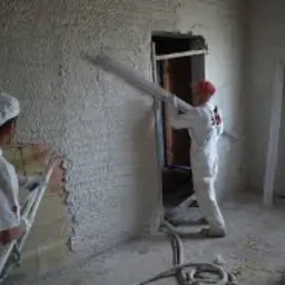 Dwóch pracowników w białych kombinezonach aplikuje tynk na ścianę w jasnym pomieszczeniu. Widoczne narzędzia i fragment okna.