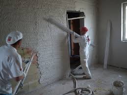 Dwóch pracowników w białych kombinezonach aplikuje tynk na ścianę w jasnym pomieszczeniu. Widoczne narzędzia i fragment okna.