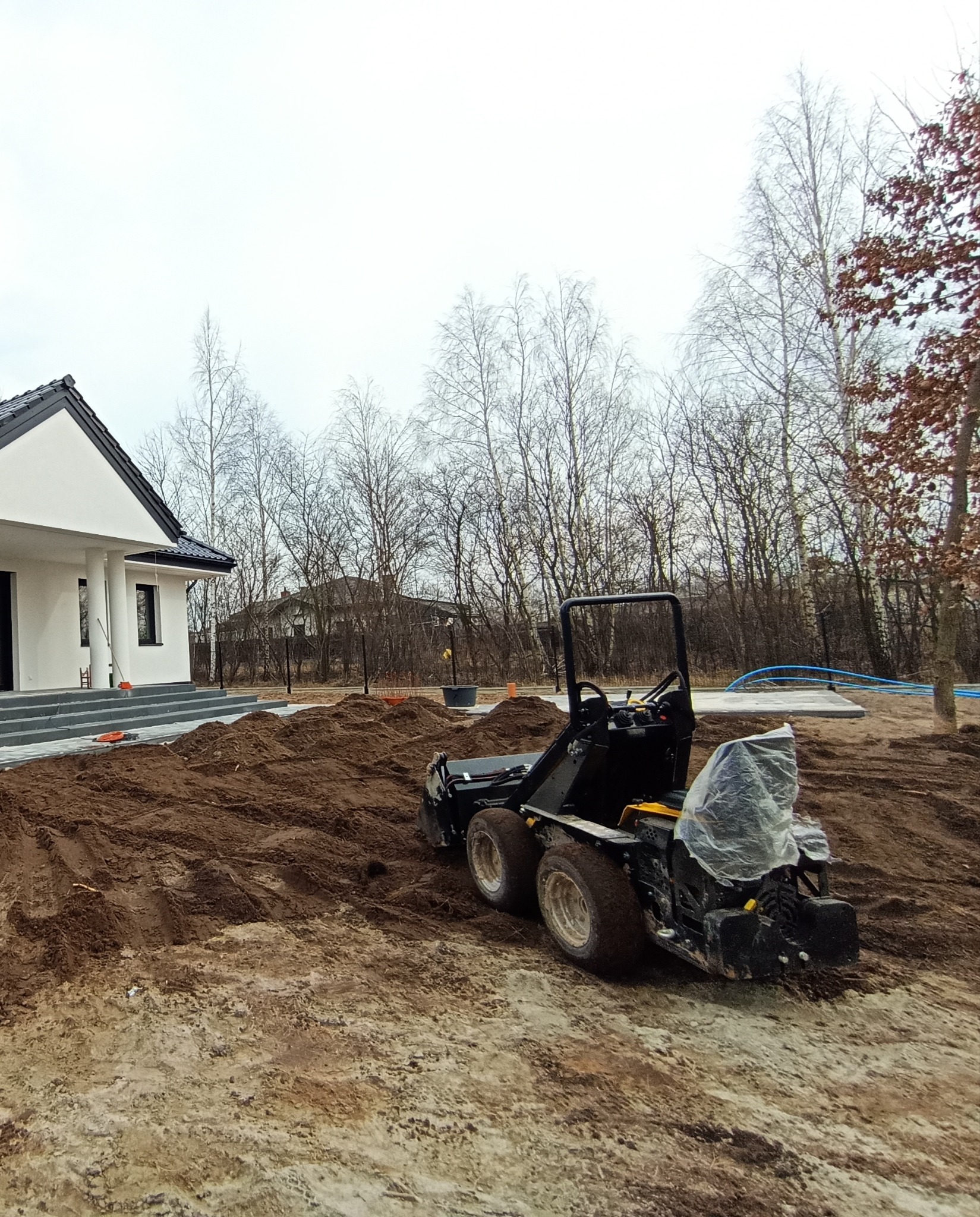 Miniładowarka na placu budowy ogrodu przed domem z białymi ścianami i szarymi schodami. W tle drzewa bez liści i błękitne rury.