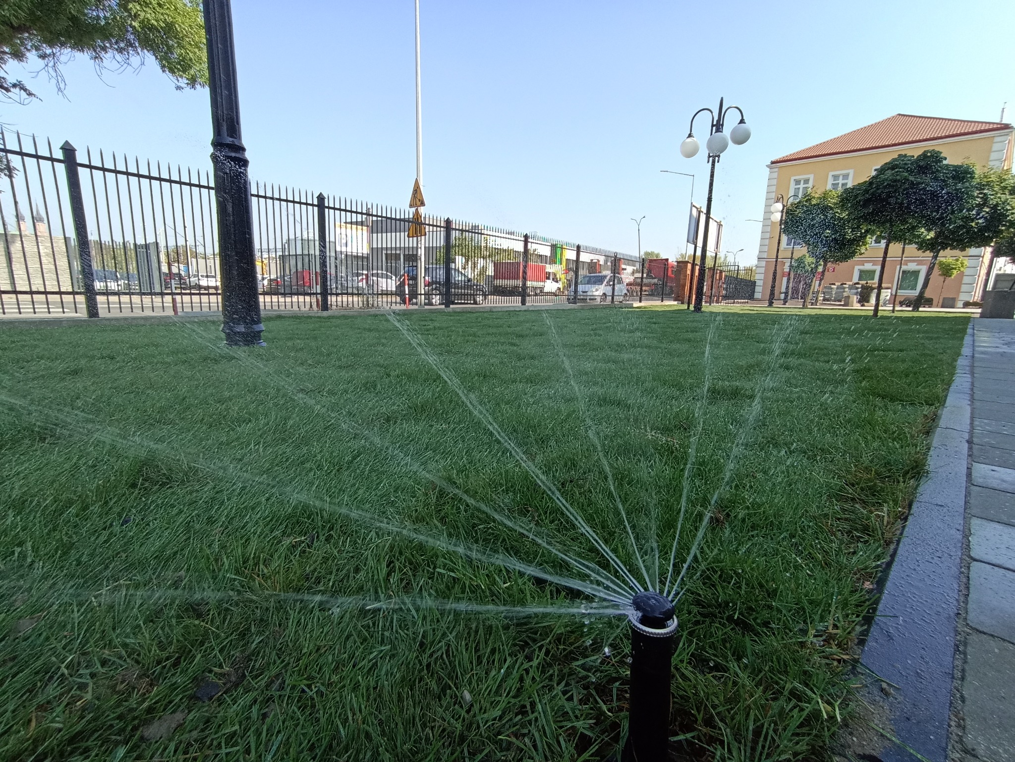 Automatyczny system nawadniania trawnika w akcji, zraszacz rozpyla wodę na tle ogrodzenia, lamp i budynku. Świeżo skoszona trawa.