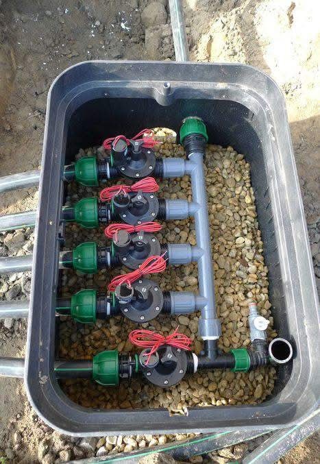 Skrzynka zaworowa z systemem nawadniania, wypełniona drobnymi kamieniami. Widoczne czarne zawory z zielonymi elementami i czerwonymi przewodami, połączone szarymi rurami.