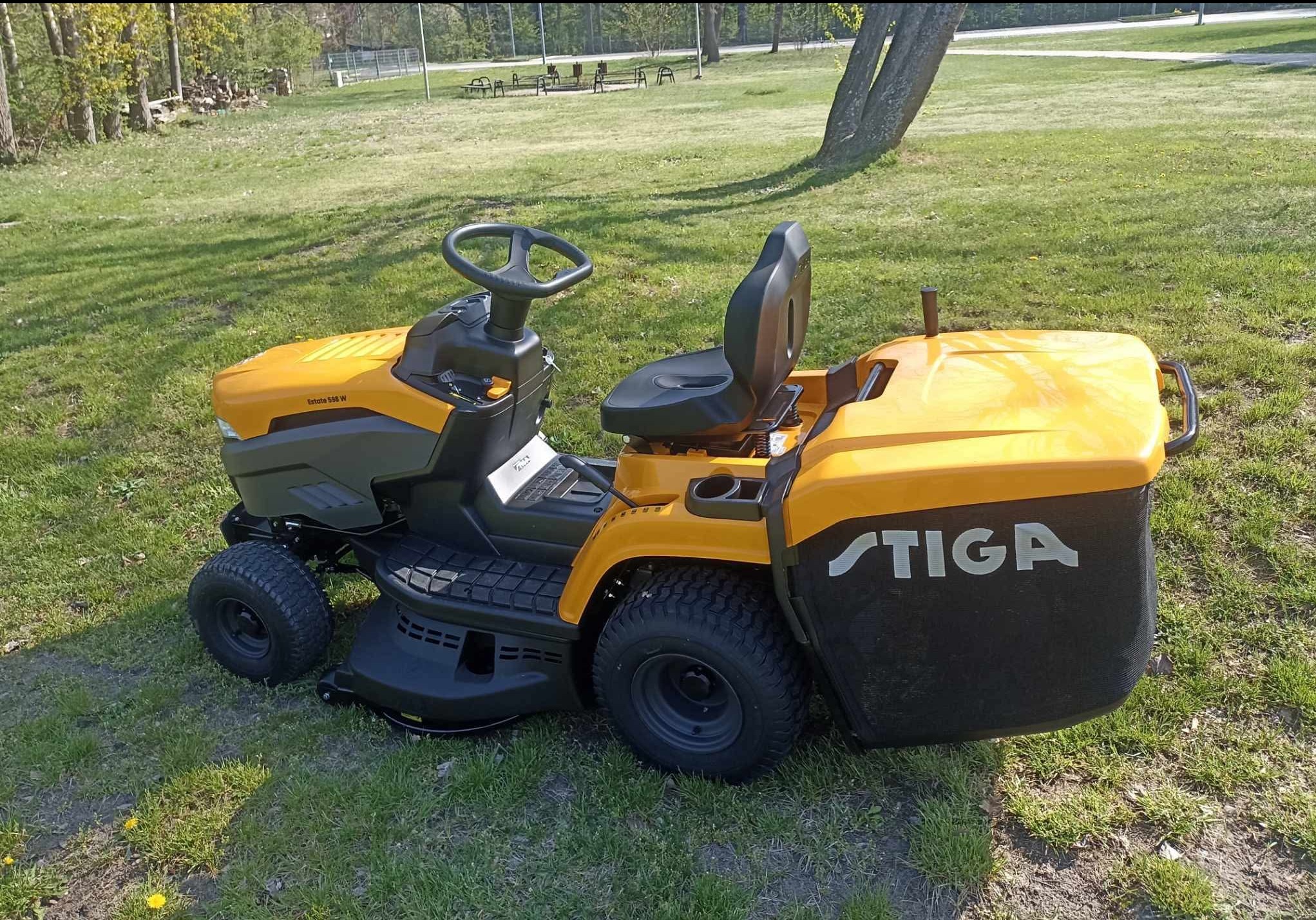 Żółto-czarny traktorek do koszenia trawy marki Stiga na zielonym trawniku, z widocznym koszem na trawę i elementami sterującymi. W tle drzewa i ławki parkowe.