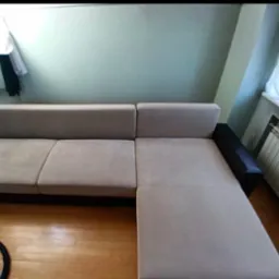 Szara, narożna kanapa z ciemnymi bokami, widoczna na drewnianej podłodze, z fragmentem węża do prania tapicerki w lewym dolnym rogu.