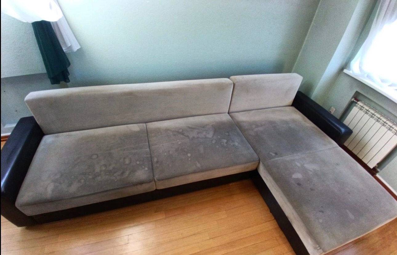 Narożnik z jasną tapicerką, widoczne plamy i zabrudzenia na siedzisku, drewniana podłoga, grzejnik w tle.