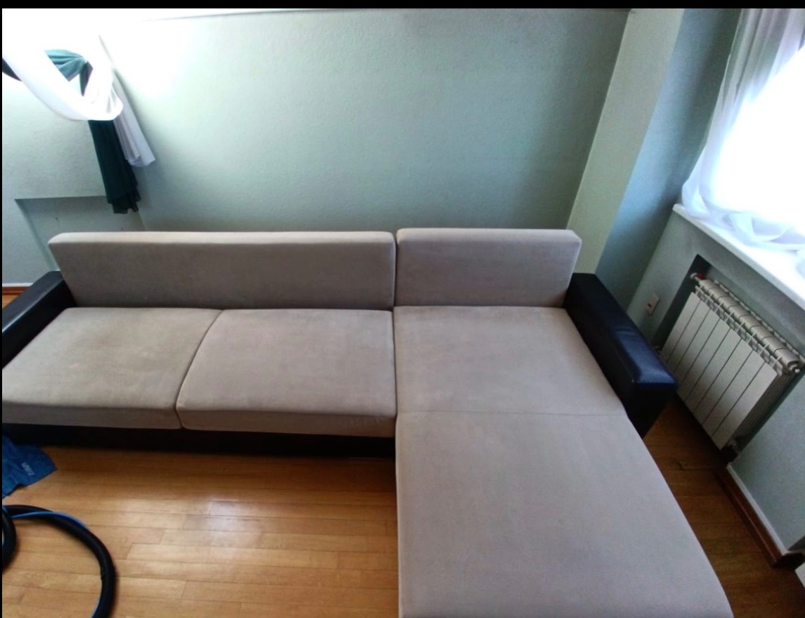 Szara, narożna kanapa z ciemnymi bokami, widoczna na drewnianej podłodze, z fragmentem węża do prania tapicerki w lewym dolnym rogu.