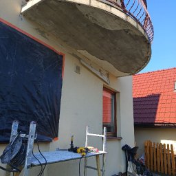 Al'kinshpul SERHII TYKHONENKO - Elewacja budynku z balkonem w trakcie prac. Widoczne rusztowanie, narzędzia i zabezpieczenia.  W tle dom z czerwoną dachówką i błękitne niebo.