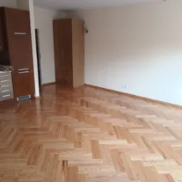 Jasny parkiet ułożony w jodełkę w pustym pokoju z brązowymi meblami kuchennymi i szafą. Świeżo wycyklinowana podłoga z widocznym połyskiem.