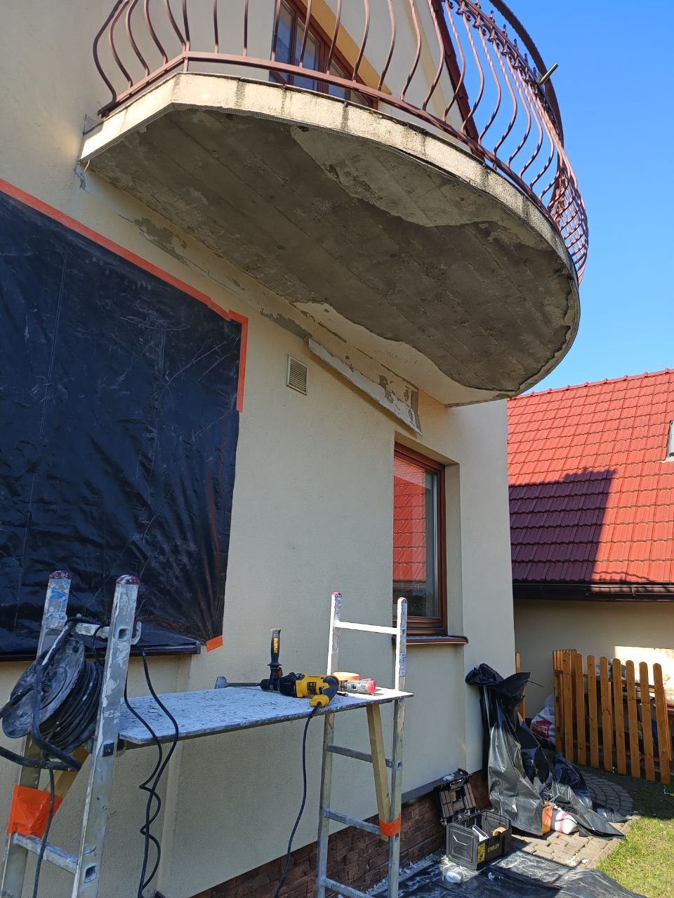 Elewacja budynku z balkonem w trakcie prac. Widoczne rusztowanie, narzędzia i zabezpieczenia.  W tle dom z czerwoną dachówką i błękitne niebo.