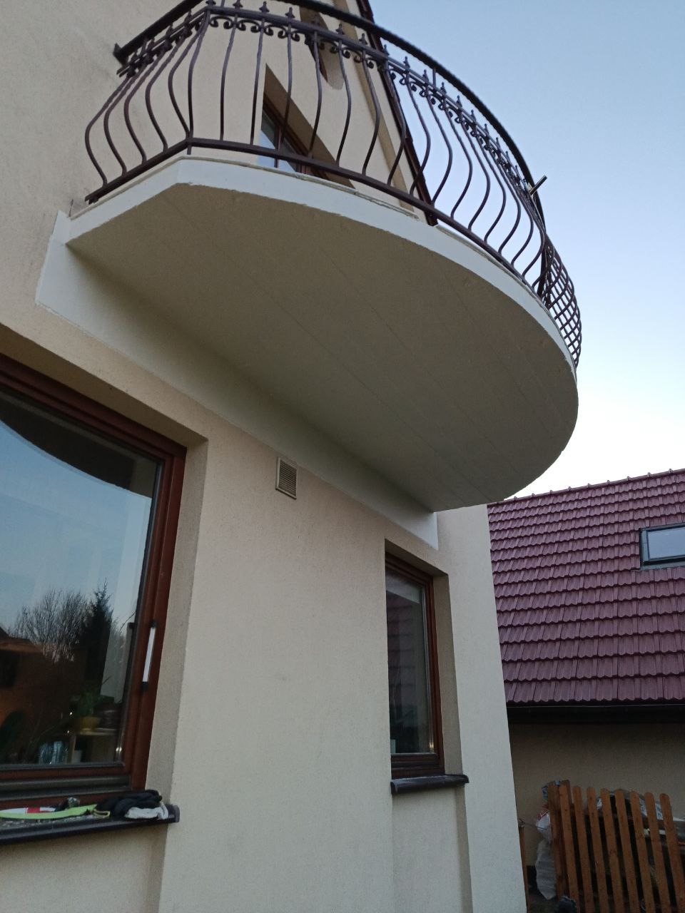Elewacja budynku z półokrągłym balkonem z ozdobną, metalową balustradą. Widoczne okna z brązowymi ramami i fragment dachu sąsiedniego budynku.