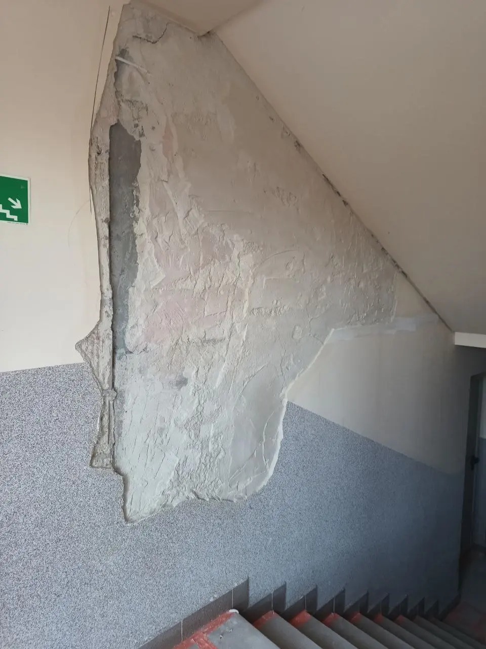 Ściana klatki schodowej w trakcie naprawy, z widocznym uszkodzeniem tynku i świeżo nałożoną warstwą gładzi. Fragment schodów w dolnej części kadru.