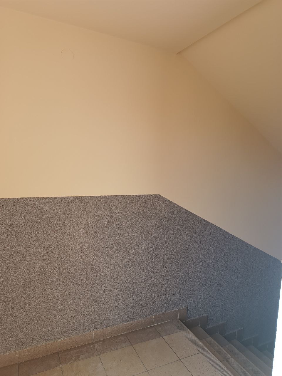 Szare schody z mozaikową ścianą i kremowym sufitem. Widok z dołu ukazuje detale wykończenia i oświetlenie. Minimalistyczny design.